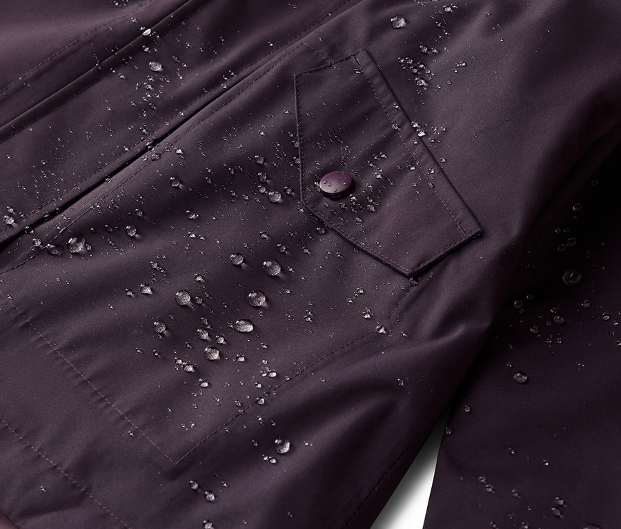 Nahaufnahme einer wasserabweisenden, auberginefarbenen Jacke mit einer Pattentasche, die mit einem Knopf geschlossen wird. Wassertropfen sind sichtbar.