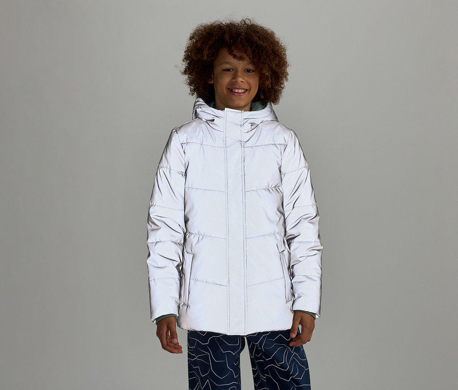 Ein lächelnder Junge mit lockigem Haar trägt eine reflektierende Pufferjacke mit Kapuze und blaue Hosen mit Muster vor einem grauen Hintergrund.
