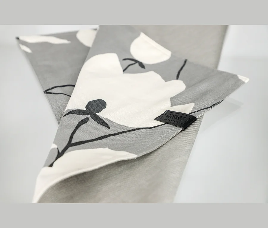 Gefaltetes graues Tischset mit weißem und schwarzem Blumenmuster und einem schwarzen Etikett der SCHÖNER WOHNEN Kollektion.