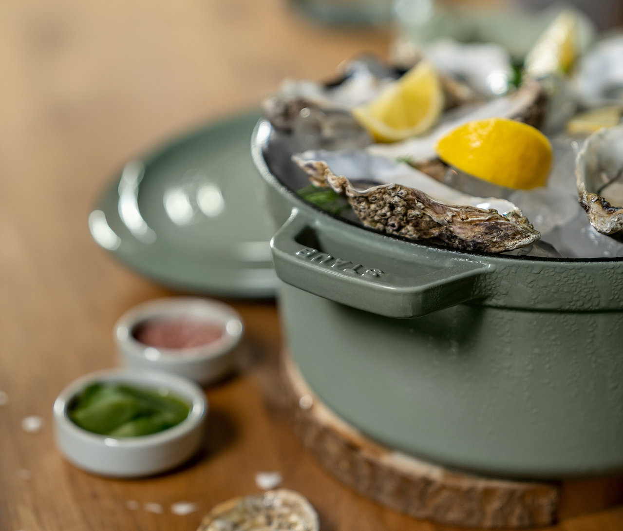 Ein grüner Topf gefüllt mit frischen Austern auf Eis, garniert mit Zitronenspalten. Daneben stehen zwei kleine Schalen mit grüner und roter Sauce auf einer Holzoberfläche.