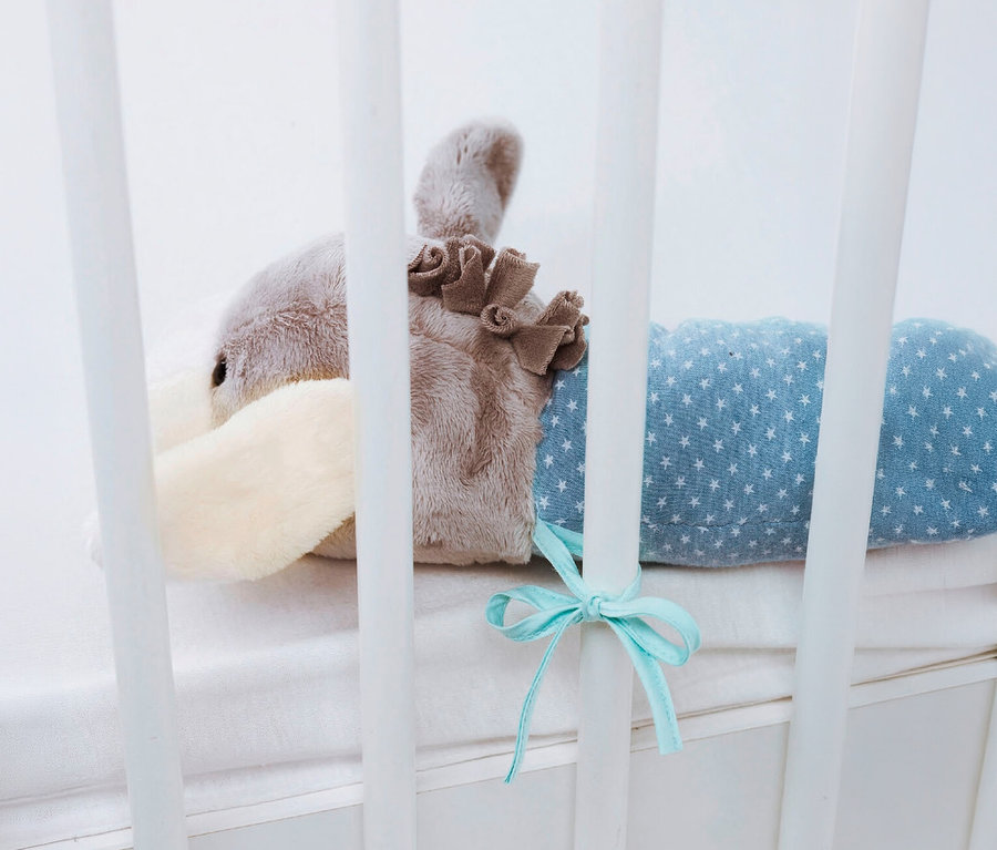 Plüschtier in Form eines grauen und cremefarbenen Esels mit blauem, sternenverziertem Körper, an den weißen Gitterstäben eines Babybetts befestigt.