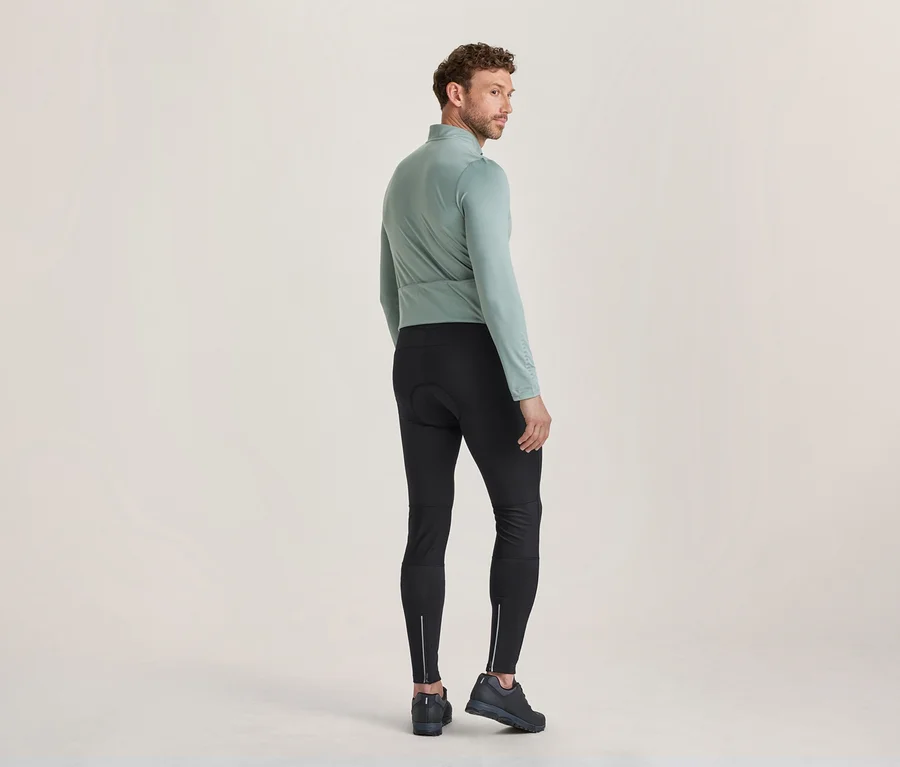 Mann steht mit dem Rücken zum Betrachter, blickt über seine rechte Schulter, trägt ein hellgrünes Langarmshirt, schwarze Radlerhosen mit reflektierenden Reißverschlüssen an den Knöcheln und dunkle Schuhe.