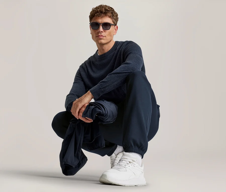 Mann mit lockigem braunem Haar und Sonnenbrille hockt, trägt ein dunkelblaues Langarmshirt, dunkelblaue Jogginghose und weiße Sneaker, hält ein dunkelblaues Kleidungsstück.