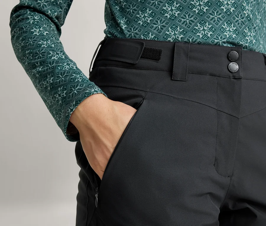 Person trägt ein grünes Langarmshirt mit geometrischem Muster und schwarze Hose, eine Hand steckt in einer Reißverschlusstasche.