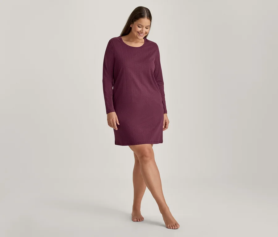 Eine lächelnde Frau in einem kurzen, langärmeligen, weinroten Kleid steht barfuß und blickt nach unten.