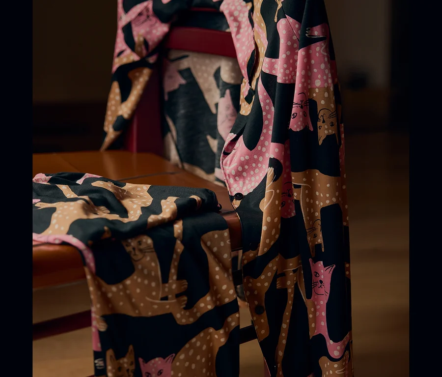 Ein schwarz, rosa und braun gemusterter Stoff mit Katzenmotiven und weißen Punkten, über einen braunen Holzstuhl drapiert.