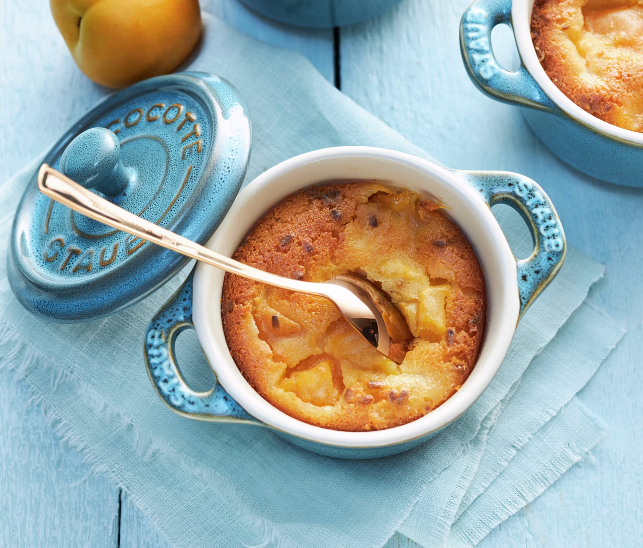 Ein goldener Löffel steckt in einem gebackenen Dessert, möglicherweise Aprikose, in einer antiktürkisen STAUB Keramik-Cocotte. Der Deckel der Cocotte liegt daneben auf einer hellblauen Serviette, mit einer weiteren Cocotte und einer gelben Frucht im Hintergrund.
