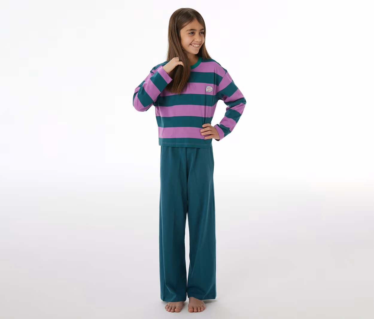 Lächelndes Mädchen in einem SCHIESSER Kinder-Pyjama mit lila-grün gestreiftem Langarmshirt und petrolfarbener Hose, barfuß.