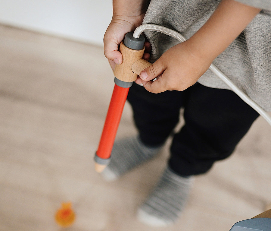 Kind hält eine rote Spielzeug-Feuerwehrspritze und zielt auf kleine orangefarbene Flammen auf dem Boden. small foot Feuerwehrrucksack.