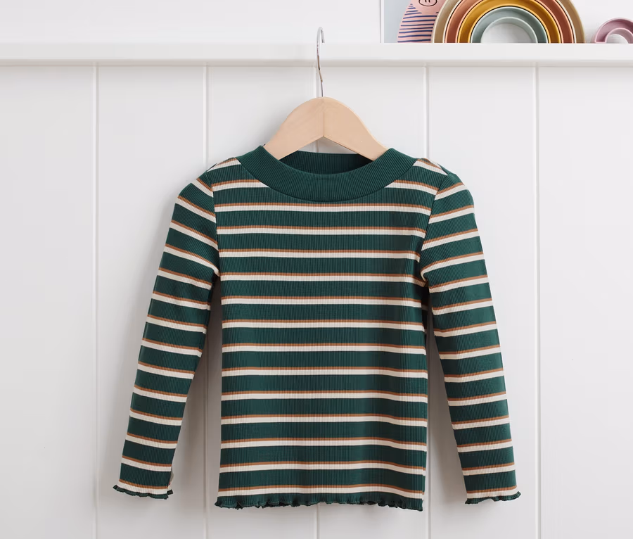Ein grün geringeltes Rippshirt hängt auf einem Bügel vor einer weißen Wand.