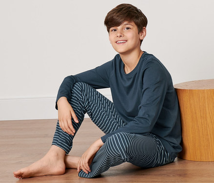Junge sitzt auf dem Boden neben einem Holzhocker und trägt einen blauen Kinder-Pyjama.