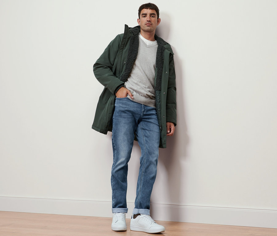 Mann steht an einer Wand, trägt einen grauen Pullover mit V-Ausschnitt, Jeans und eine Parka.