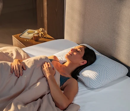 Frau schläft friedlich auf dem Rücken im Bett, zugedeckt mit einer hellen Bettdecke, auf einem weißen, konturierten TEMPUR® ARCH SmartCool™ Schlafkissen mit blauem Muster. Sonnenlicht fällt auf ihr Gesicht.