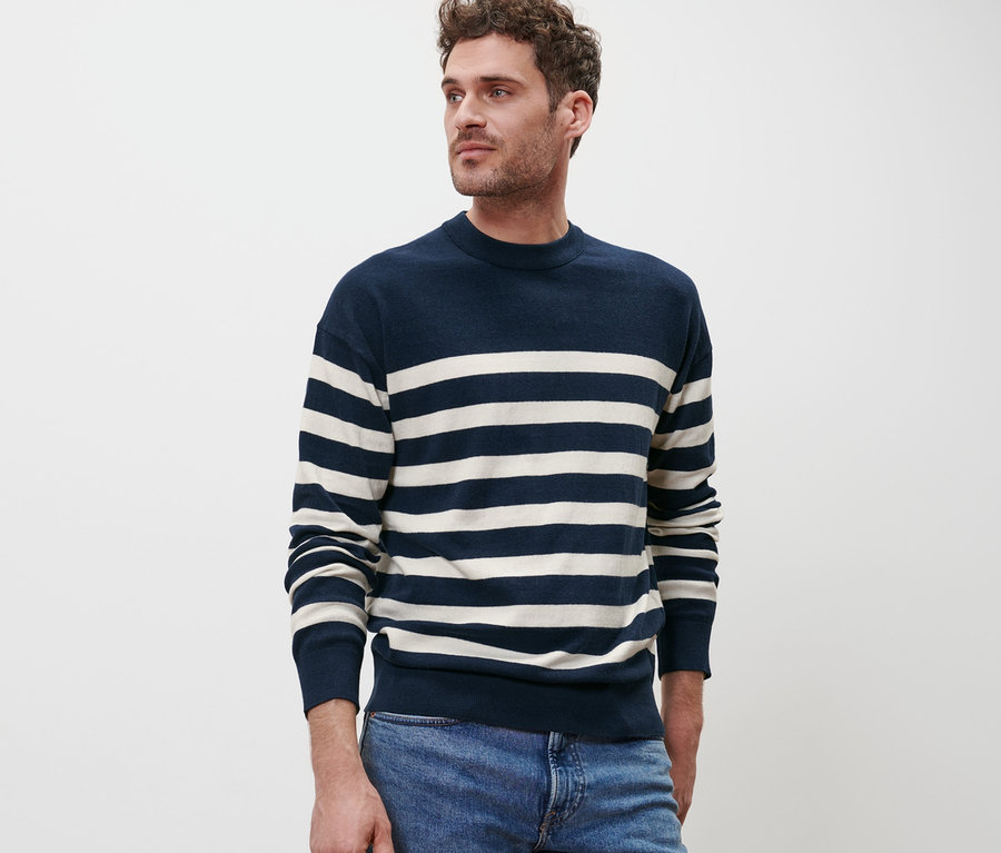 Mann posiert vor einem weißen Hintergrund in einem Pullover mit Rundhalsausschnitt mit blau-weißen Streifen und blauen Jeans.