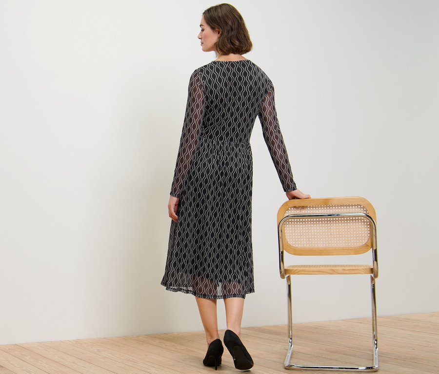 Frau von hinten in einem bedruckten Mesh-Kleid, die Hand auf einem Stuhl.