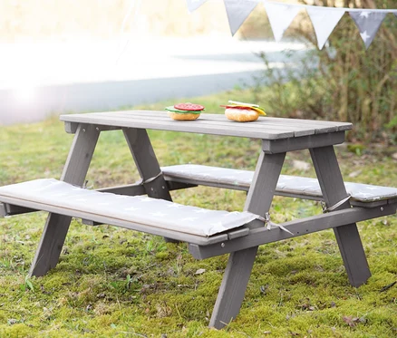Eine graue Kinder-Outdoor-Sitzgruppe "Picknick for 4" von Roba steht auf einer Wiese. Auf dem Tisch liegen zwei Spielzeughamburger.