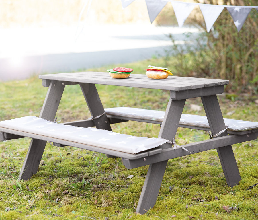 Eine graue Kinder-Outdoor-Sitzgruppe "Picknick for 4" von Roba steht auf einer Wiese. Auf dem Tisch liegen zwei Spielzeughamburger.