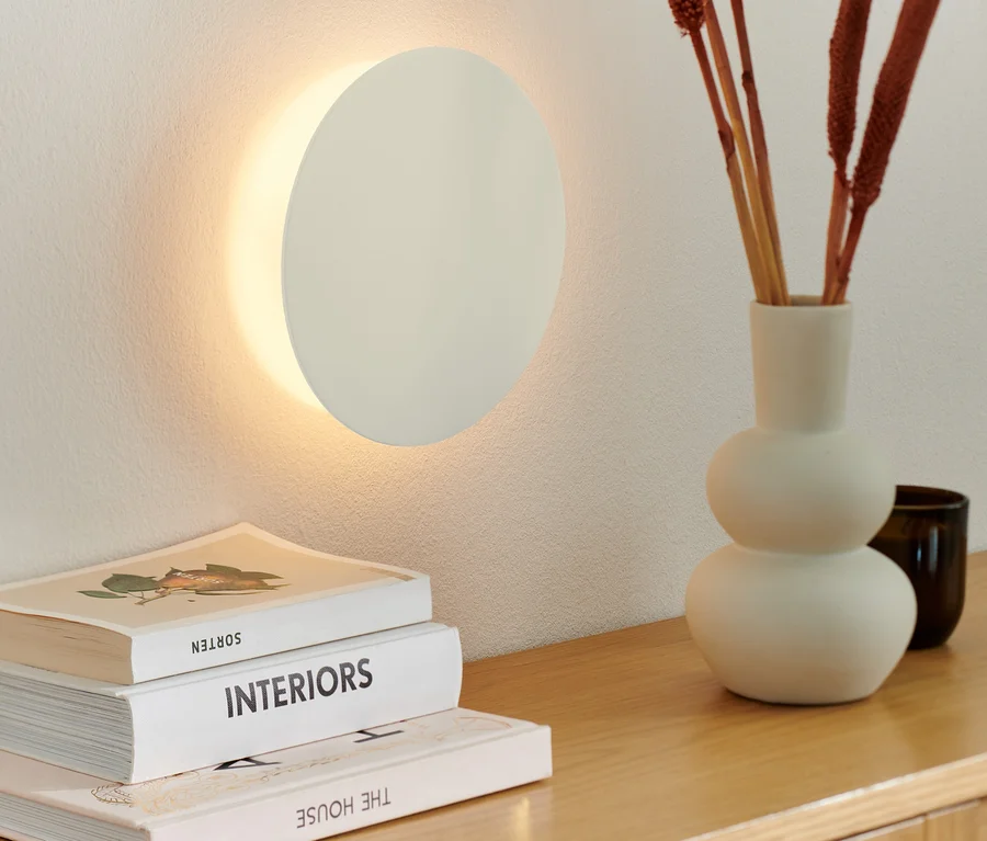 An der Wand ist eine LED-Akku-Wandleuchte montiert. Auf einer Holzkommode liegen eine Vase mit Zierästen und Bücher.