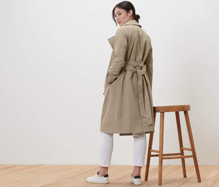 Frau posiert in beigem Trenchcoat und weißen Hosen neben einem Holzhocker.