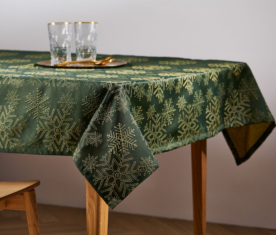 Auf einem Holztisch liegt eine grüne Jacquard-Tischdecke mit Schneeflockenmuster. Auf dem Tisch stehen Gläser und ein Holzstuhl.