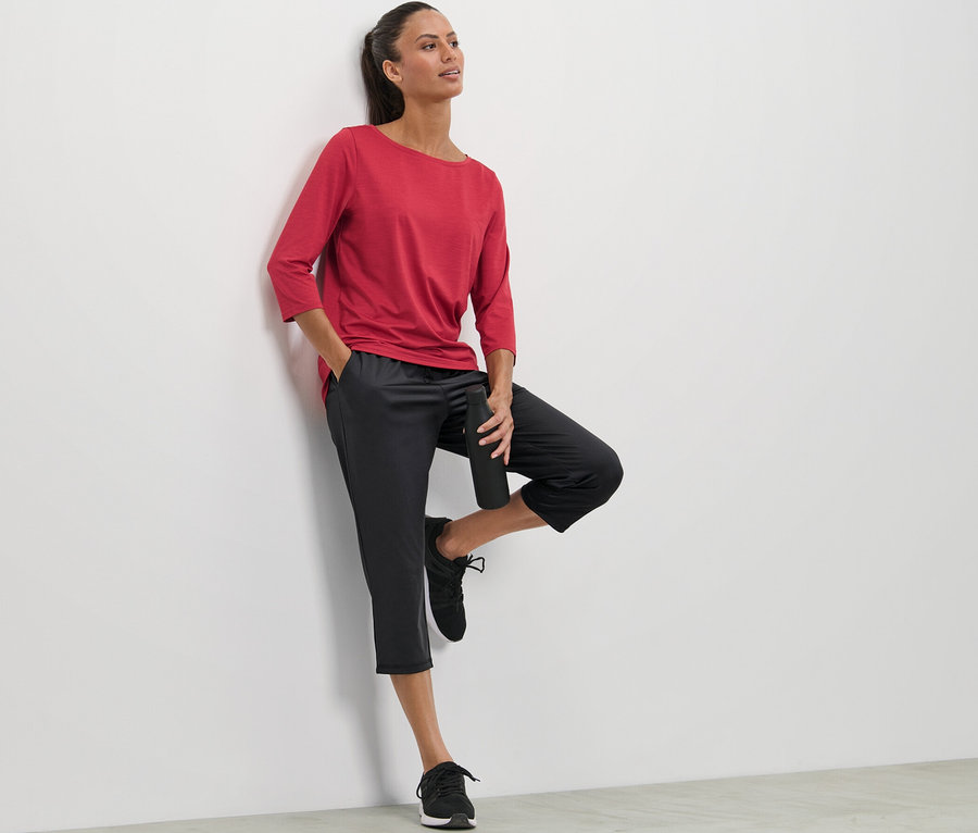 Eine Frau lehnt an einer Wand und trägt ein rotes T-Shirt und eine 3/4-Sporthose.