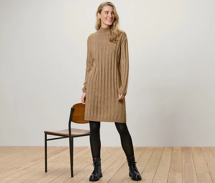 Eine Frau steht neben einem Stuhl und trägt ein geripptes Strickkleid mit Stehkragen.