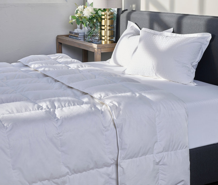 Eine gesteppte, weiße Home By TEMPUR® Luxe Duo-Decke liegt auf einem Bett mit grauem Kopfteil.