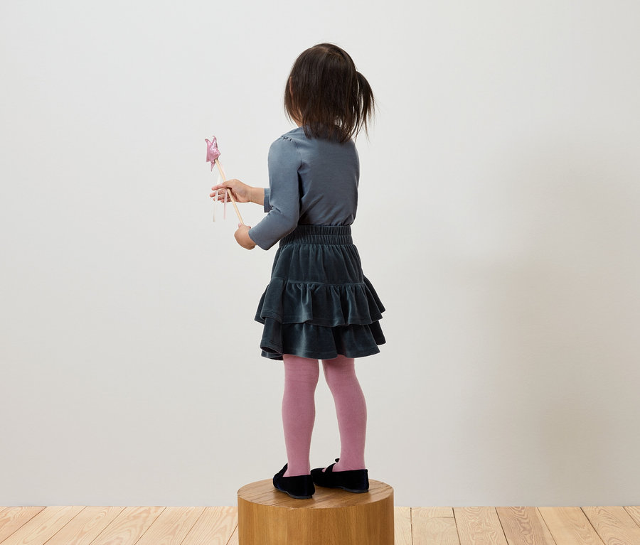 Mädchen in grauem Shirt und grünem Rock steht auf einem Holzsockel und hält einen Zauberstab.