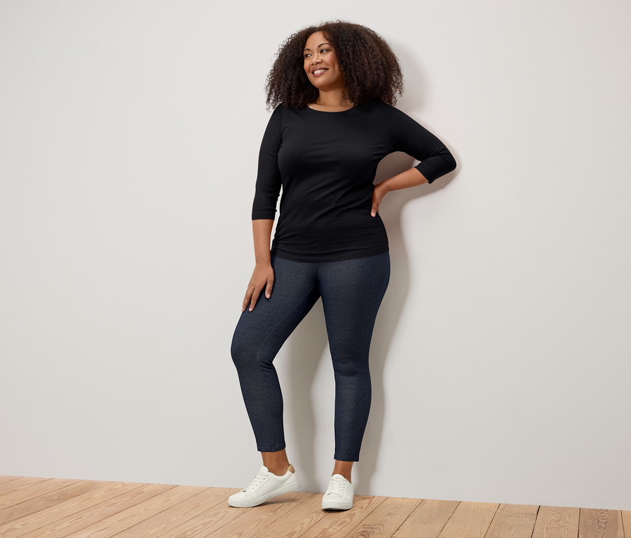 Lächelnde Frau posiert in schwarzem Dreiviertelarm-Shirt, dunkelblauen Jeggings und weißen Sneakers.