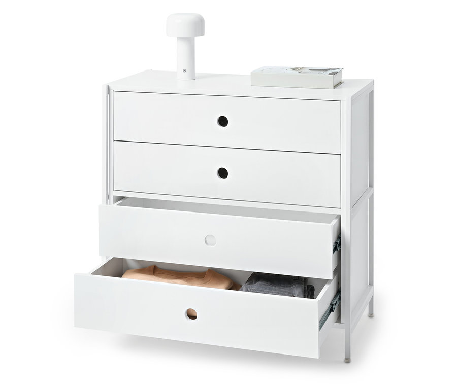 Weisse Kommode mit drei Schubladen, von denen zwei offen sind und Kleidung enthalten. Auf der Kommode stehen eine Lampe und ein Buch.