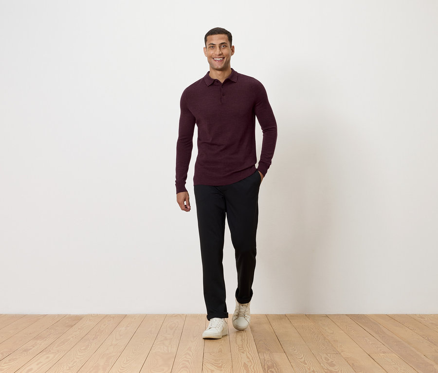 Mann geht auf Holzboden. Er trägt ein weinrotes langärmeliges Poloshirt, schwarze Hosen und weiße Turnschuhe.