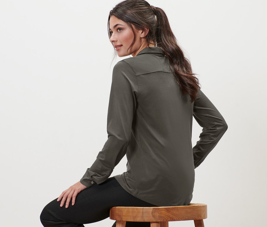 Frau sitzt auf einem Holzhocker und trägt ein Blusenshirt mit Knopfleiste.