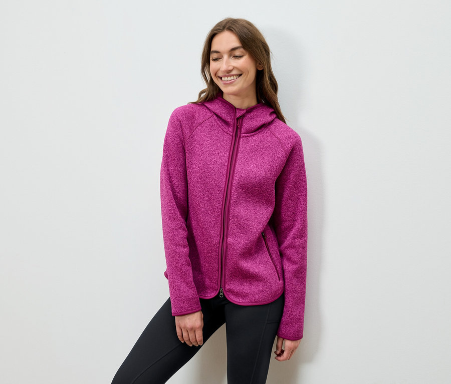 Lächelnde Frau posiert in einem pinkfarbenen Kapuzenpullover und schwarzen Hosen.