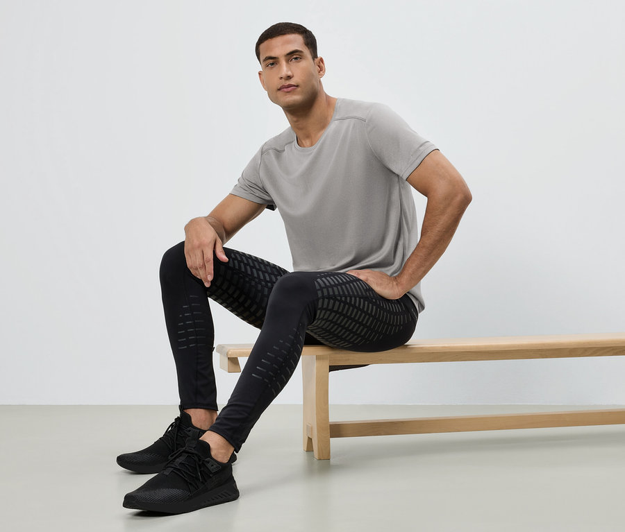 Mann sitzt auf einer Bank, trägt ein graues T-Shirt, schwarze Leggings mit Muster und schwarze Turnschuhe.