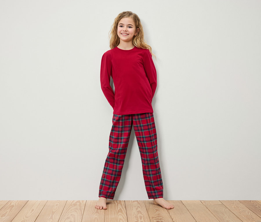 Mädchen im roten Langarm-Pyjama und karierter Hose steht auf einem Holzboden vor einer weißen Wand.