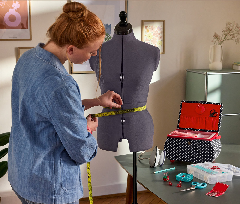 Frau misst die Taille an der Prym Multi-Schneiderbüste »Prymadonna« ab. Daneben: Prym Nähkorb »Polka Dots«, Prym Mini-Dampfbügeleisen, Prym Starter-Set Nähen »Love«.