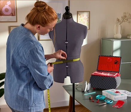 Frau misst die Taille an der Prym Multi-Schneiderbüste »Prymadonna« ab. Daneben: Prym Nähkorb »Polka Dots«, Prym Mini-Dampfbügeleisen, Prym Starter-Set Nähen »Love«.