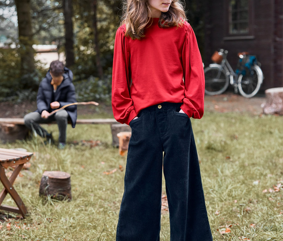Mädchen mit Cord-Culotte und Langarmshirt steht draussen mit Händen in den Hosentaschen.