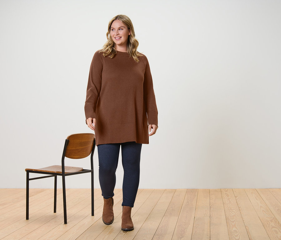 Eine Frau trägt einen langen, dunkelbraunen Strickpullover und gefütterte Leggings in Jeansoptik und geht.