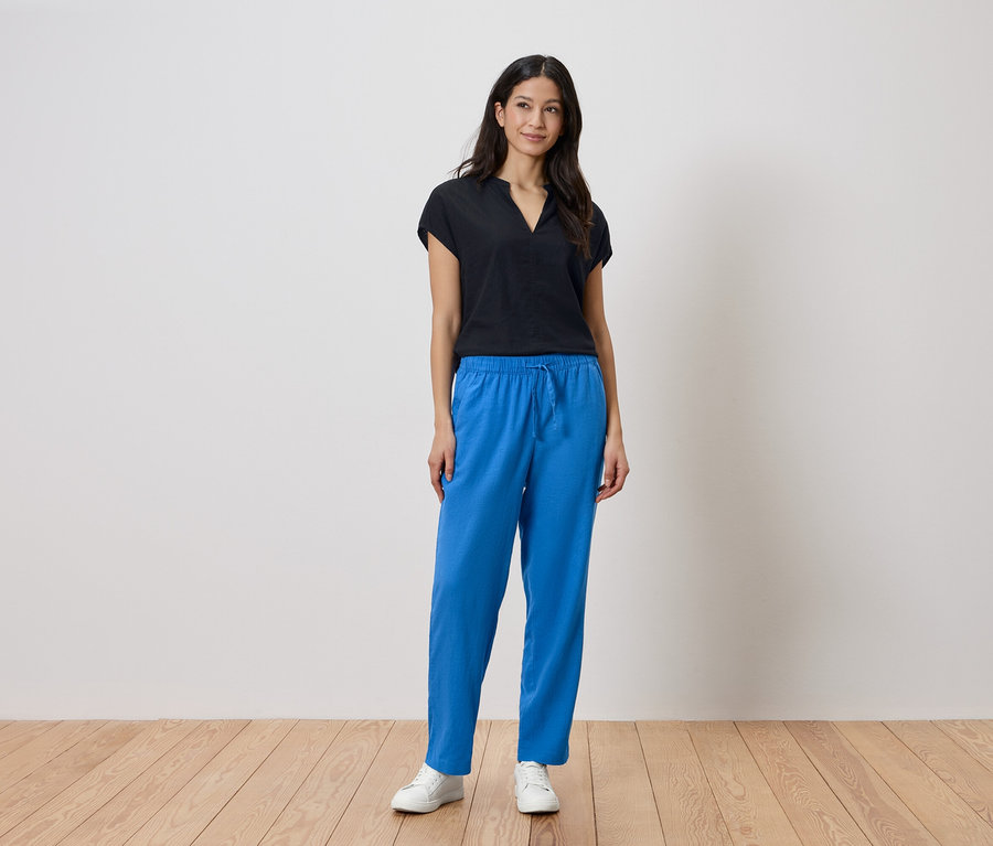 Frau steht in schwarzem Oberteil und blauen Hosen vor einer Wand.