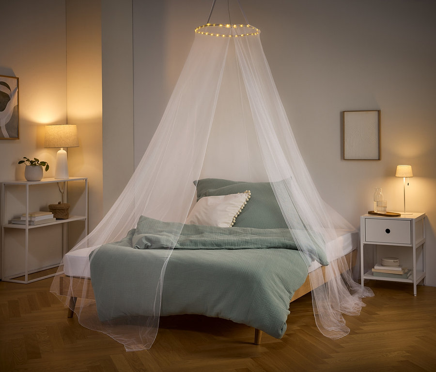 Ein Bett mit grüner Bettwäsche und einem weißen Moskitonetz mit Lichtern hängt von der Decke. Nachttisch und Regal stehen an den Seiten.