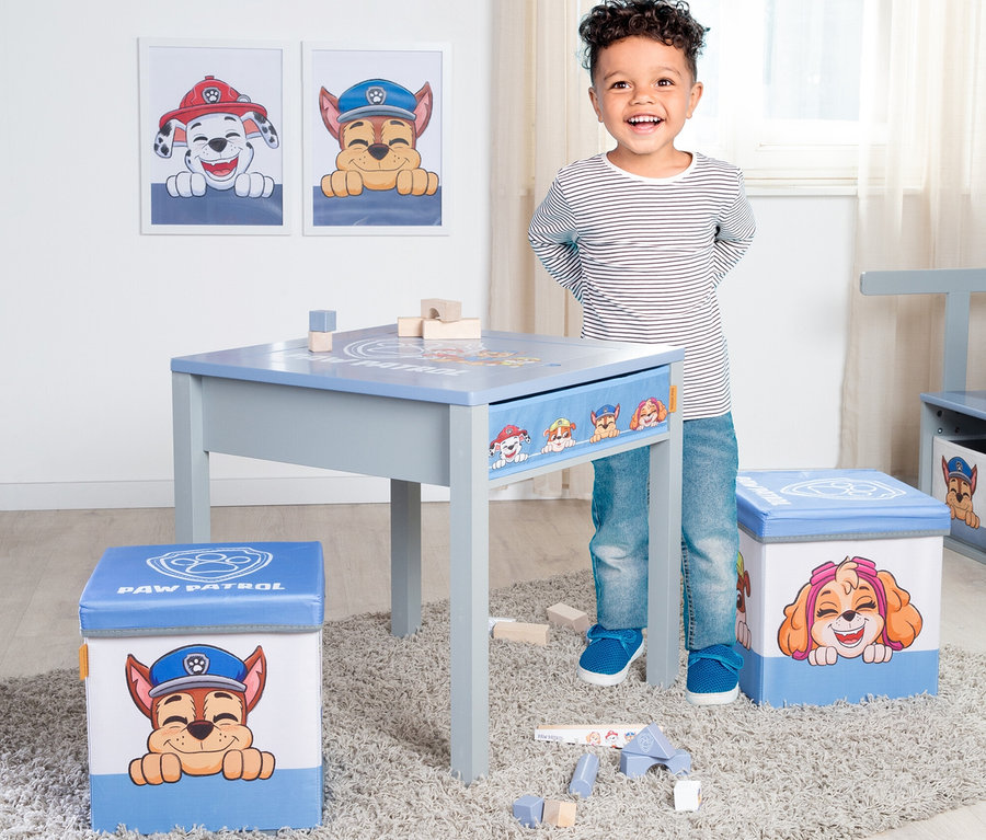 Ein lachender Junge steht neben einem grauen Kindertisch mit Paw Patrol-Motiven und zwei Paw Patrol-Hocker stehen auf einem Teppich.