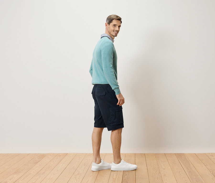 Ein Mann steht seitlich, mit Blick über die Schulter, und trägt einen hellblauen Pullover, kurze, dunkle Cargohosen und weiße Turnschuhe.