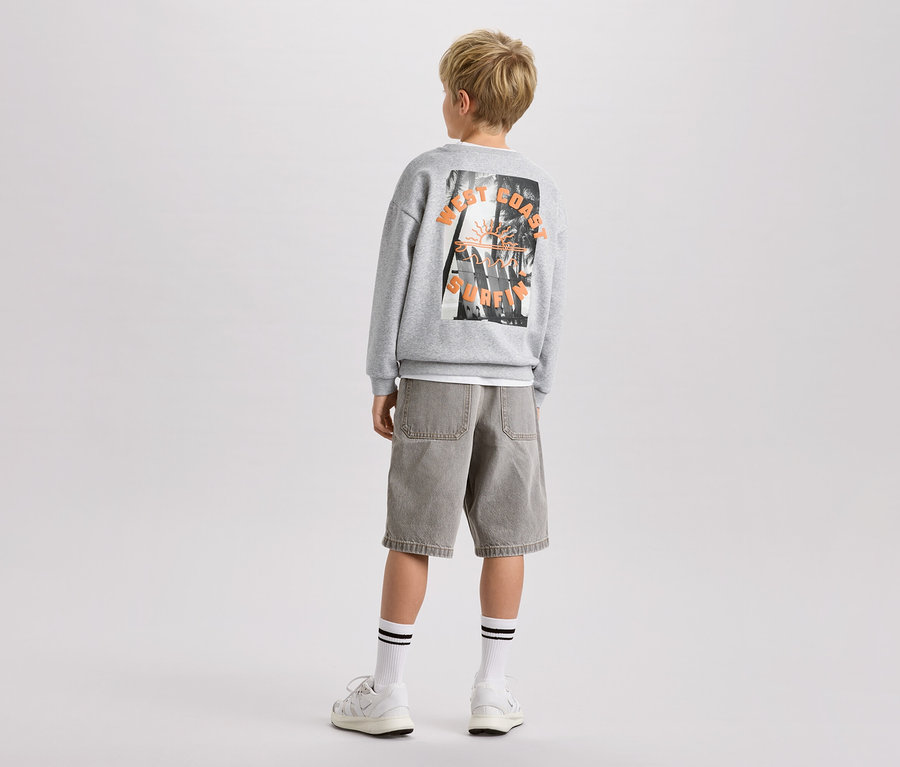 Junge von hinten, trägt graues Sweatshirt mit Aufdruck, Shorts und Sportschuhe.