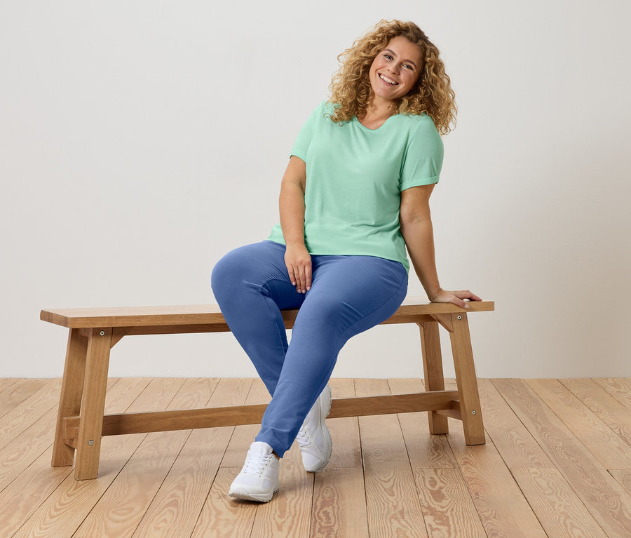 Eine Frau mit lockigem Haar sitzt auf einer Holzbank, trägt ein grünes T-Shirt und blaue Hosen.