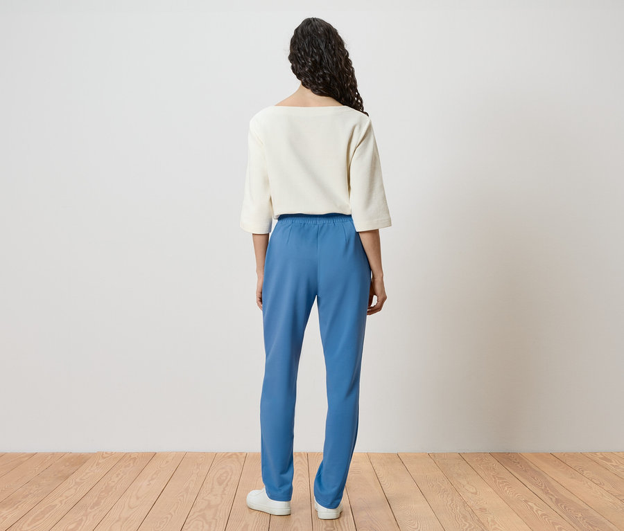 Rückansicht einer Frau mit lockigem Haar, die ein weißes Oberteil, blaue Hosen und weiße Schuhe trägt.