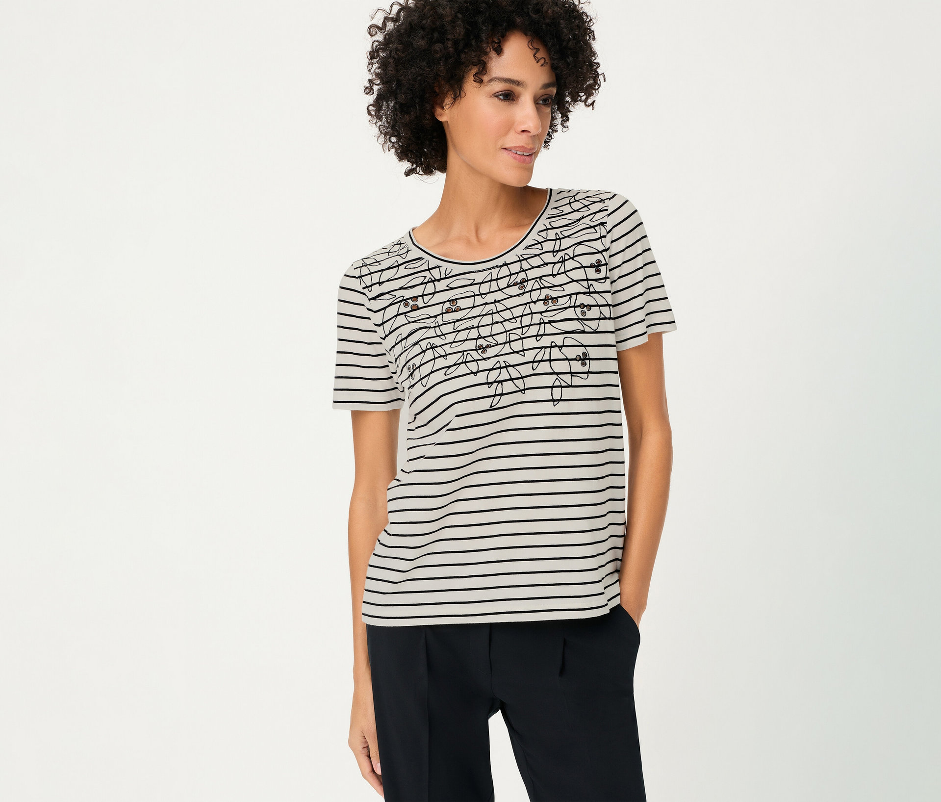 Frau mit lockigem Haar trägt ein gestreiftes Olsen Shirt mit Blumenmuster und dunkle Hose.