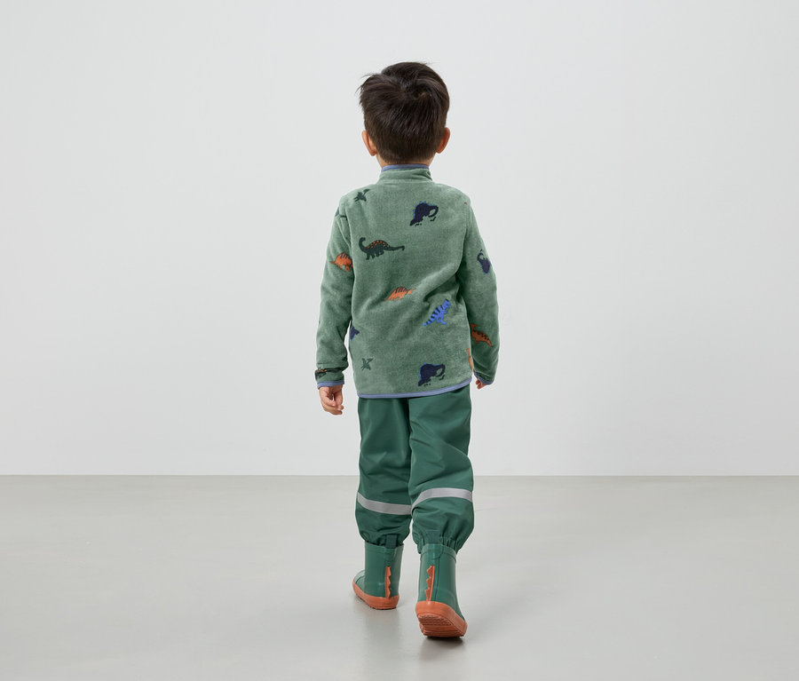 Ein Kind von hinten, trägt einen grünen Fleecepullover mit Dinosauriern, grüne Regenhose und Gummistiefel.