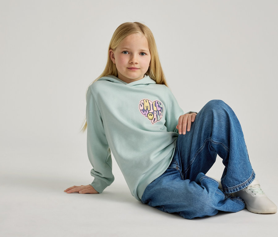 Ein blondes Mädchen sitzt auf dem Boden und trägt einen hellblauen Hoodie mit einem Smiley-Herz-Aufdruck und weite Jeans.