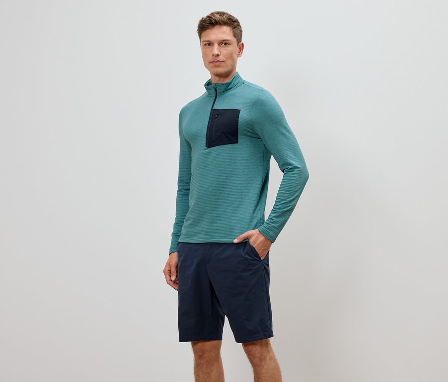 Mann mit türkisfarbenem Langarmshirt mit Reißverschluss und marineblauen Shorts, eine Hand in der Hosentasche.